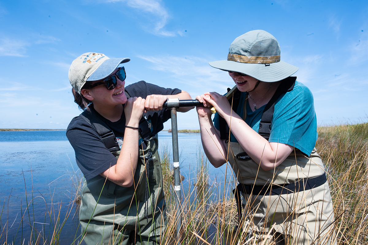 OBXFS_Currituck_FW_Sept-30 Two women in waders laugh as they attempt to dislodge a soil core from the marsh.