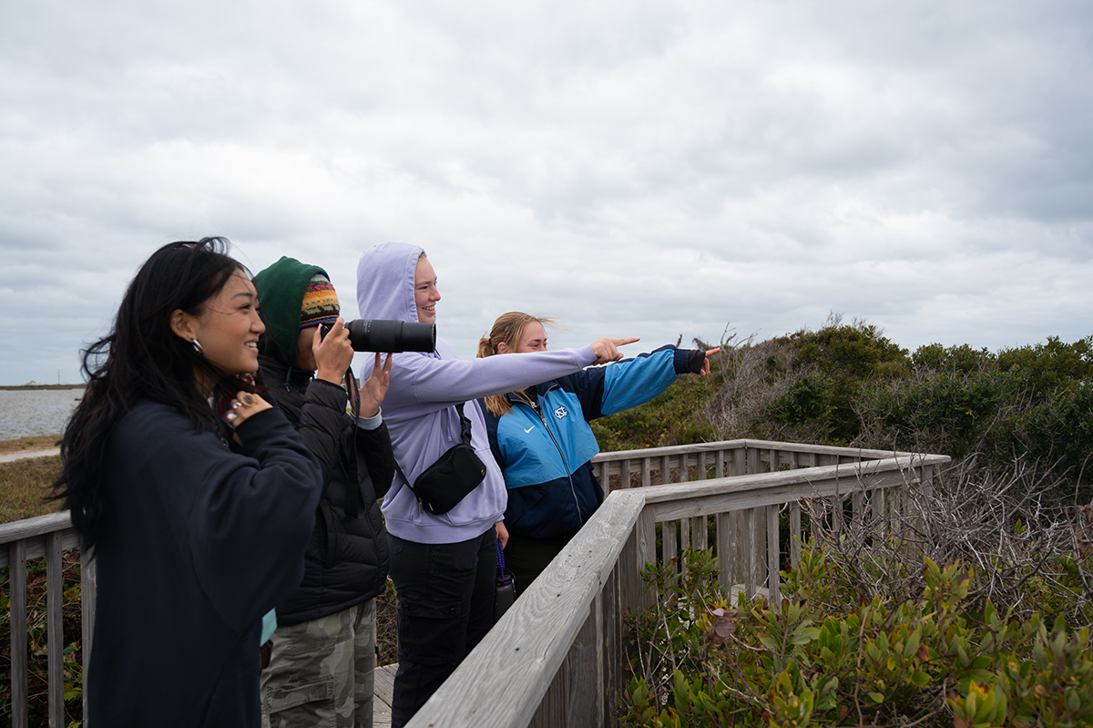 OBXFS_Rodanthe-28 On an overcast day, four girls stand on a boardwalk with binoculars and cameras in hand. They point at something not seen in the image.
