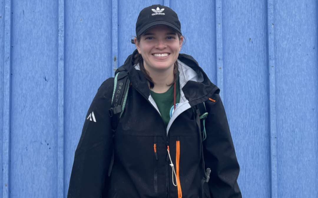 A woman wearing a ball cap and cold weather gear stands in front of a blue building and smiles.
